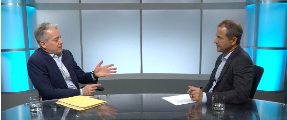 Two men in business attire are seated at a glass table in a modern studio setting, engaged in a serious conversation. One man gestures with his hand while holding a pen, and the other listens attentively while holding a piece of paper. Both have glasses of water in front of them, and the background features illuminated blue panels.