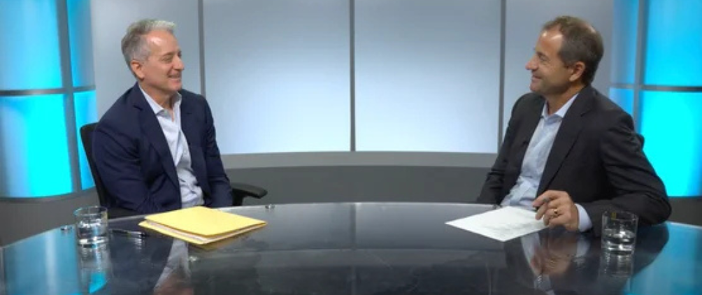 Two men in business attire sit across from each other at a glass table, engaged in conversation. Both are smiling, with papers and glasses of water in front of them. A modern studio background with blue lighting is visible.