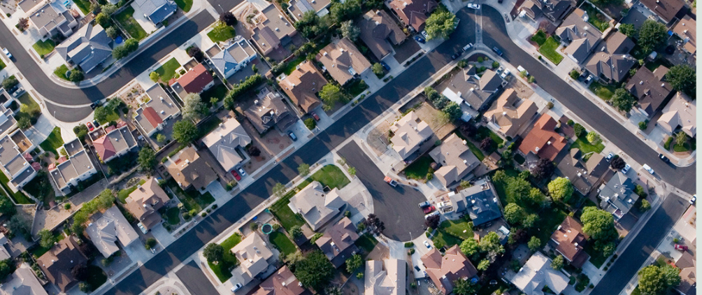 Aerial view of a suburban neighborhood with rows of houses, tree-lined streets, driveways, and backyards, showing a grid-like street layout.