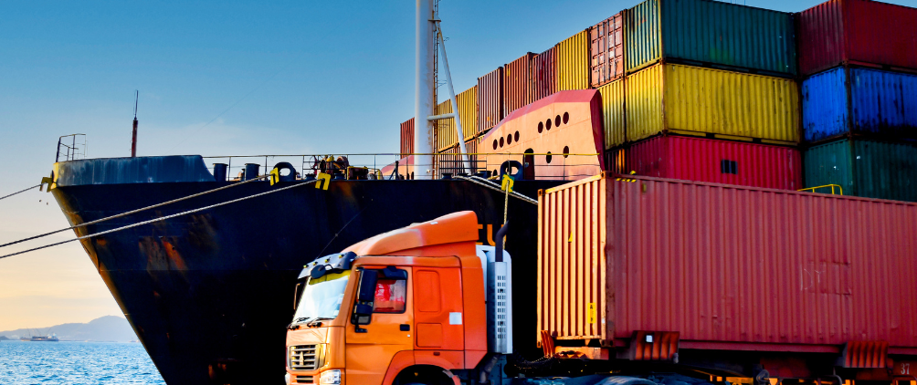 Cargo ship docked at a port with stacked colorful shipping containers and an orange truck carrying a container in the foreground