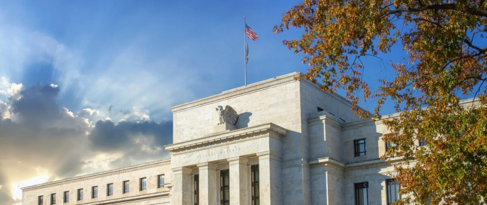 The Federal Reserve building in Washington, D.C., with the U.S. flag flying on top, sunlight streaming through clouds in the background, and autumn tree branches on the right.